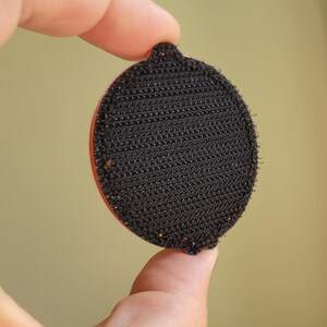 May include: A round, black hook-and-loop fastener with a red edge is held between fingers. The fastener has a textured surface and a small tab at the top. The background is a solid, muted yellow-green.
