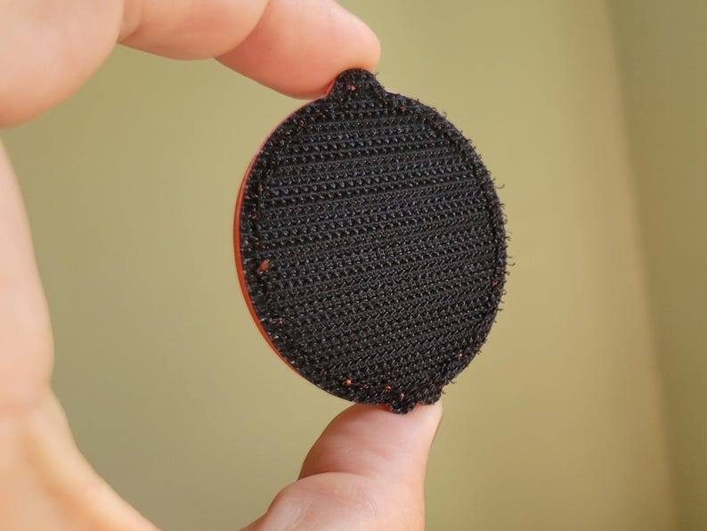 May include: A round, black hook-and-loop fastener with a red edge is held between fingers. The fastener has a textured surface and a small tab at the top. The background is a solid, muted yellow-green.