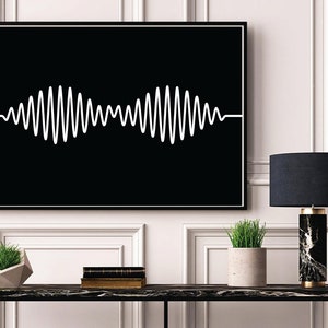 May include: A black and white framed print of a white waveform on a black background. The print is hanging on a white wall above a black console table with a black and white marble lamp, a potted plant, and a stack of books.