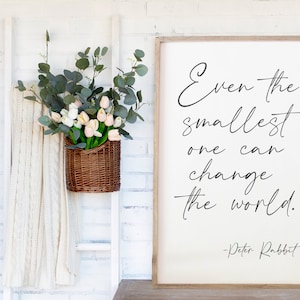 May include: A white framed sign with the quote "Even the smallest one can change the world." attributed to Peter Rabbit. The sign is hanging on a white brick wall with a wicker basket filled with pink and white flowers and greenery hanging on a white ladder.
