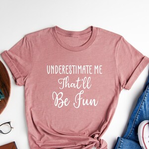 May include: A heather mauve t-shirt with the text "UNDERESTIMATE ME That'll Be Fun" in white lettering. The shirt is shown on a white background with a pair of jeans, white trainers, a wooden tray with a plant, a camera, and glasses.