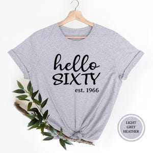 A grey t-shirt with the text "Hello SIXTY est. 1966" printed on it, along with a wooden hanger. The t-shirt is displayed against a white background, and there is a branch of green leaves next to it.