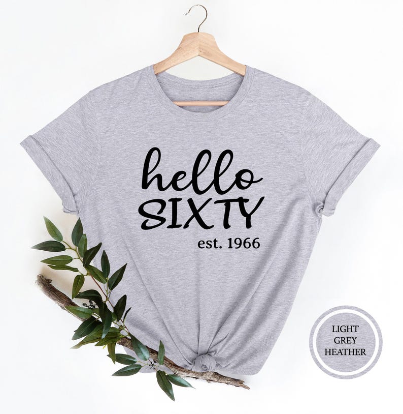 A grey t-shirt with the text "Hello SIXTY est. 1966" printed on it, along with a wooden hanger. The t-shirt is displayed against a white background, and there is a branch of green leaves next to it.