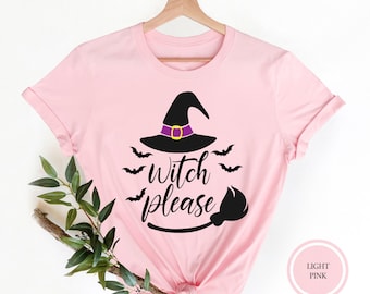 May include: A light pink t-shirt featuring a black witch hat with a purple band, a black broomstick, and several small black bats. The text "Witch please" is printed in a black cursive script in the center of the graphic design.