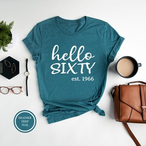 A teal t-shirt with the text "Hello SIXTY est. 1966" printed on it, along with a brown leather handbag, a watch, a pair of glasses, and a small plant.