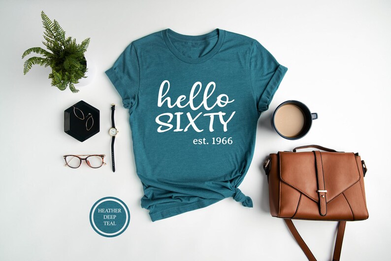A teal t-shirt with the text "Hello SIXTY est. 1966" printed on it, along with a brown leather handbag, a watch, a pair of glasses, and a small plant.