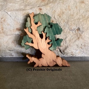 May include: A wooden tree-shaped ornament with a light brown trunk and green leaves. The tree is layered, with the trunk and branches in a lighter shade than the leaves. The text "(C) Peachie Originals" is at the bottom.
