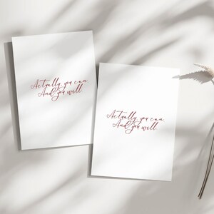May include: Two white cards with the text "Actually, you can. And you will" in a burgundy script font. The cards are on a white surface with shadows and a dried flower stem.