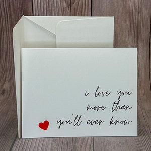 Op de afbeelding: Een witte kaart met een rood hart en de tekst "i love you more than you'll ever know" in zwarte cursieve letters.