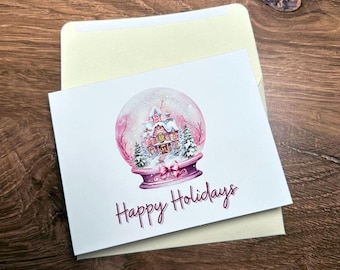 May include: A white greeting card with a watercolor illustration of a snow globe featuring a house and trees. The snow globe is set on a purple base with a bow. The words "Happy Holidays" are written in a cursive font below the snow globe. The card is inside a cream-colored envelope.