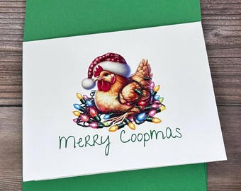 May include: A white card with a green envelope. The card features a cartoon illustration of a chicken wearing a Santa hat and surrounded by Christmas lights. The text "Merry Coopmas" is written in green cursive.