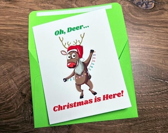 May include: A Christmas greeting card with a cartoon reindeer wearing a Santa hat and holding string lights. The card reads "Oh, Deer... Christmas is Here!" and is inside a bright green envelope.