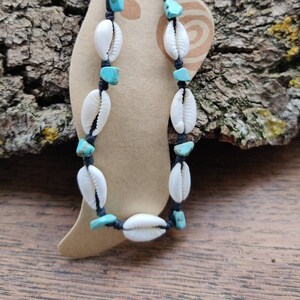 Peut inclure: Deux chevillères avec des coquillages blancs et des perles turquoise sur un cordon noir. Les chevillères sont présentées sur une étiquette en papier brun avec le texte "Beach Style" imprimé dessus.