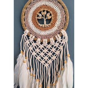 Può includere: Un acchiappasogni fatto a mano con un design circolare. Il centro presenta un albero fatto di conchiglie. L'anello esterno è tessuto con filato marrone, beige e bianco. Piume bianche e perline di legno pendono dal basso.