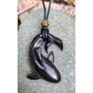 Puede incluir: Colgante de tiburón de madera marrón oscuro, suspendido de un collar de cordón negro. El tiburón está tallado en una pose curvada, con aletas y cola detalladas. Una pequeña cuenta dorada adorna el cordón sobre el colgante. Mide unos 7 cm.