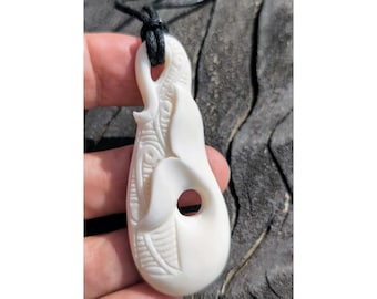 May include: A white bone pendant carved with intricate designs, featuring a hole for a cord. The pendant is hanging from a black cord.