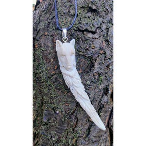 May include: A white wolf pendant carved from antler with a black cord necklace. The wolf's head is detailed with fur and the body is carved into a long, flowing tail.