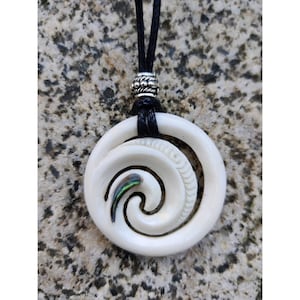 Pode incluir: Pingente de osso branco com um design em espiral e uma pequena incrustação de abalone iridescente. O pingente está em um cordão preto com uma conta de prata.
