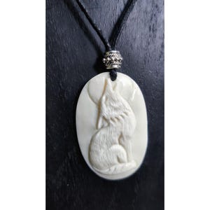 Op de afbeelding: Crème-kleurige ovale hangerketting met een gebeeldhouwd ontwerp van een huilende wolf. De hanger hangt aan een zwart koord met een decoratieve zilverkleurige kraal. De wolf is afgebeeld in een gedetailleerde, zittende pose.