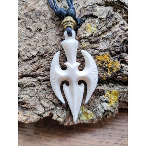 May include: White bone pendant carved in the shape of a traditional Maori tiki, with intricate details and a black cord necklace.