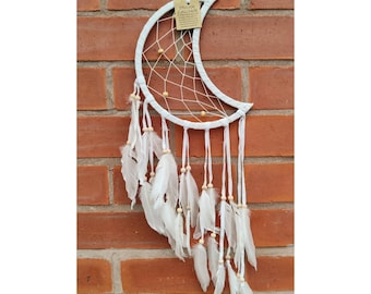 May include: White crescent moon dream catcher with a woven web and hanging white feathers. The dream catcher has small wooden beads and a tag that reads "Dream Catcher". The dream catcher is hanging against a red brick wall.