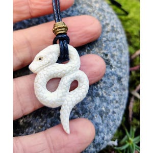May include: White bone snake pendant necklace with a black cord. The snake is carved in detail and has a realistic appearance.
