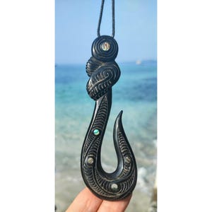 Pode incluir: Pêndulo preto esculpido em forma de anzol de pesca com incrustações de madrepérola. O pêndulo está em um cordão preto.