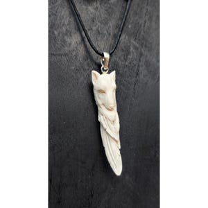 May include: A pendant featuring a carved off-white wolf head transitioning into a feather shape, suspended from a thin black cord. The wolf carving displays detailed facial features and fur texture, attached to the cord by a silver-toned metal bail.