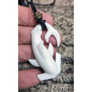 May include: Cream-coloured pendant necklace featuring a carved shark design. The shark pendant is suspended from a black cord with a decorative bronze-coloured clasp. The shark pendant is approximately 7.6 cm long.