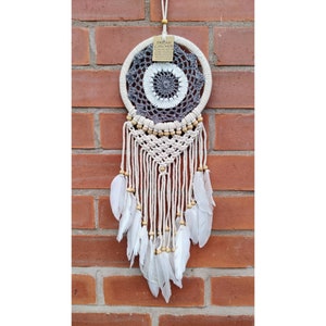 May include: A white and grey dream catcher with white feathers hanging from it. The dream catcher is hanging on a brick wall. The tag on the dream catcher says "Dream Catcher".