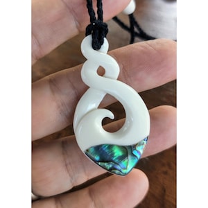 Pode incluir: Pingente de osso branco com incrustação de madrepérola verde e azul. O pingente tem a forma de um design tradicional Maori koru, um símbolo de novos começos e crescimento. Está pendurado em um cordão preto.