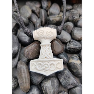 May include: White carved bone pendant of Thor's hammer, Mjolnir, with intricate details. The pendant is on a black cord and is surrounded by smooth grey stones.