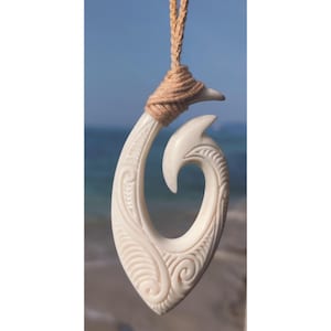 Peut inclure: Un pendentif en os blanc en forme d'hameçon de pêche avec des sculptures complexes, suspendu à un cordon brun naturel.