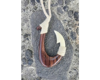 Puede incluir: Un collar con un anzuelo de pesca de madera con una punta de hueso blanco. El anzuelo es marrón con veta de madera oscura y está atado con un cordón blanco.
