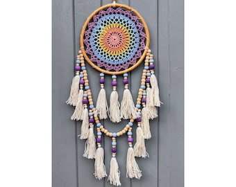 Puede incluir: Un atrapasueños artesanal con un aro de madera y un centro de ganchillo colorido en tonos rosa, naranja, amarillo, verde, azul y morado. Cuenta con borlas color crema, cuentas de madera y cuentas moradas, creando una estética bohemia.