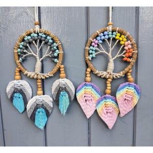 Puede incluir: Dos atrapasueños con cuentas de madera y plumas. Los atrapasueños presentan un diseño de árbol de la vida con cuentas de colores. Un atrapasueños tiene plumas azules, grises y blancas, mientras que el otro tiene plumas rosas, amarillas y naranjas.
