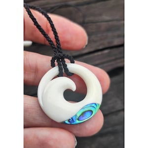 Può includere: Un ciondolo in osso bianco con un design a spirale e un'intarsio di abalone blu e verde. Il ciondolo è su un cordoncino nero.