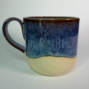 May include: A ceramic mug featuring a smooth, glossy blue glaze on the upper portion that transitions into a textured, matte beige base. The handle is finished in a dark brown glaze, matching the rim of the mug, creating a distinct contrast.