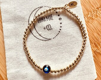 May include: Gold beaded bracelet with a blue evil eye charm. The bracelet is in a small white drawstring bag with the text "Luna Mae Co." and "LMC" printed on it.