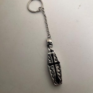 Op de afbeelding: Zilverkleurige sleutelhanger met een doodskistvormige hanger met een kruisontwerp. De hanger is bevestigd aan een ketting en een sleutelring. Een kleine schedel bevindt zich bovenop de doodskist. De sleutelhanger is waarschijnlijk van metaal.