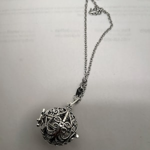 Peut inclure: Collier argenté avec un pendentif sphérique ajouré et détaillé. Le pendentif présente un design orné et complexe avec une ouverture à charnière. La chaîne est de style délicat et maillé, et le collier est présenté sur un fond neutre.
