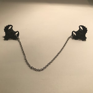May include: A pair of black metal collar clips connected by a silver chain.