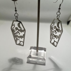 May include: A pair of silver coffin-shaped earrings with a crescent moon and floral design. The earrings are hanging from a clear acrylic earring stand.