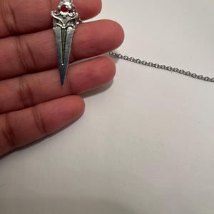 May include: A silver pendant necklace with a dagger-shaped charm featuring a red gemstone. The chain is silver and has a delicate link design.