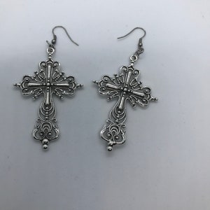 Può includere: Un paio di orecchini a croce argentati con dettagli intricati. Gli orecchini sono di stile pendente e hanno una chiusura a gancio.