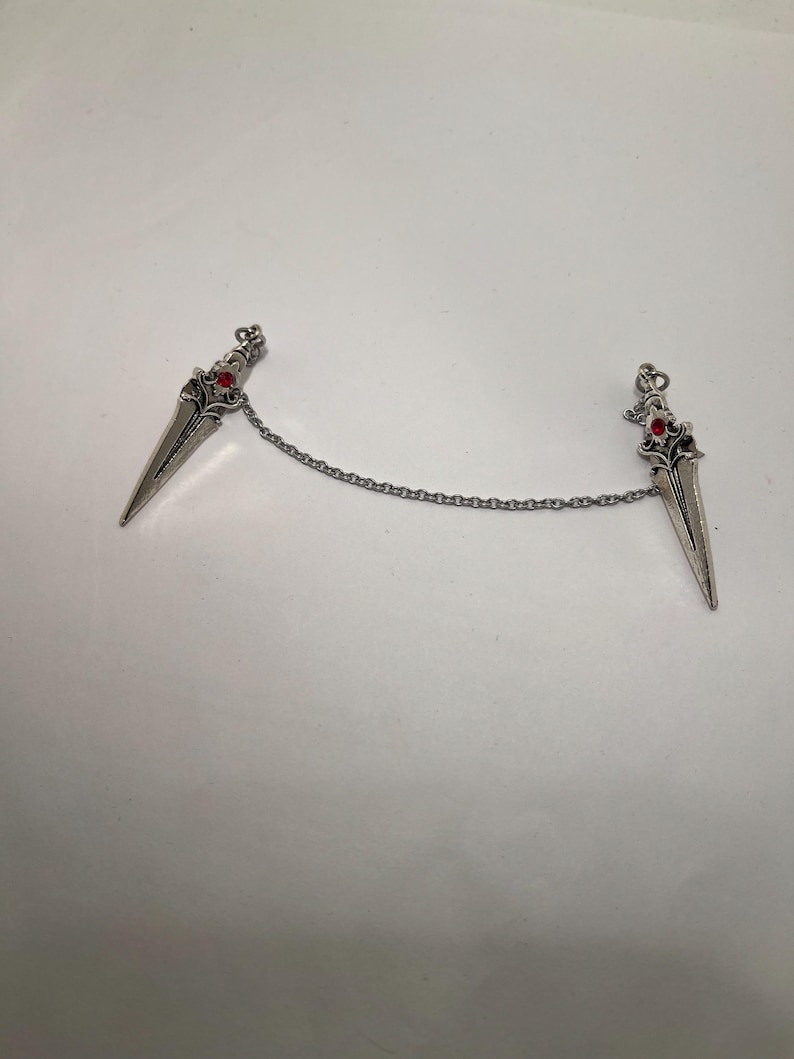 May include: A silver chain with two silver dagger charms. Each dagger has a red gemstone at the top.