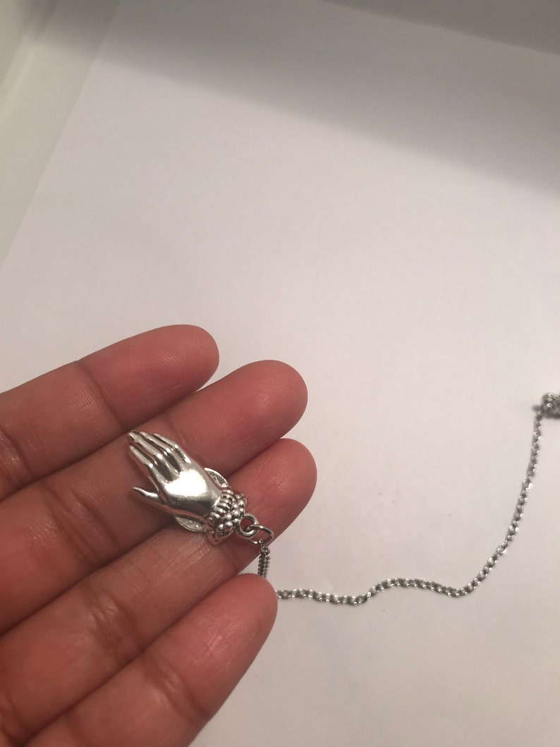 May include: A silver necklace with a hand charm. The hand is detailed with a textured bracelet.