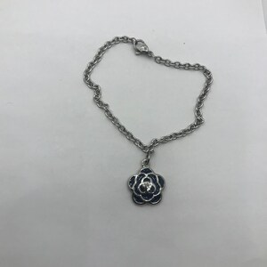 Puede incluir: Pulsera de cadena plateada con un colgante de flor. El colgante presenta una flor de esmalte azul oscuro con una pequeña gema transparente en el centro. Sobre fondo blanco.
