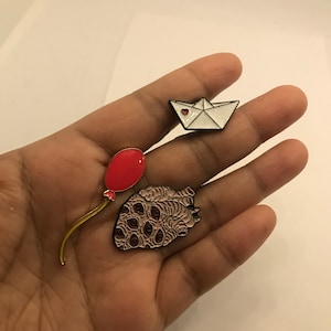 Op de afbeelding: Een verzameling emaille pins met een rode ballon, een witte papieren boot met een rood hart en een gestileerd hart met meerdere ogen. De pins hebben zwarte contouren en worden in een hand gehouden.