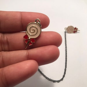 May include: A gold chain necklace with a white and red lollipop charm. The lollipop has a gold swirl design and a red bow.